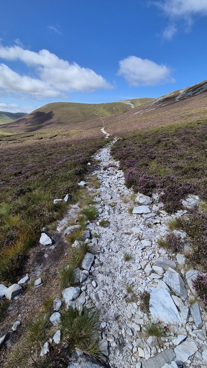The pale stony path across heather towards the hill under a blue sky.