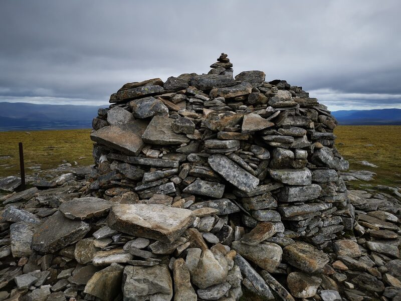 The large summit cairn of stacked slabs with an old fence post, under a grey sky.