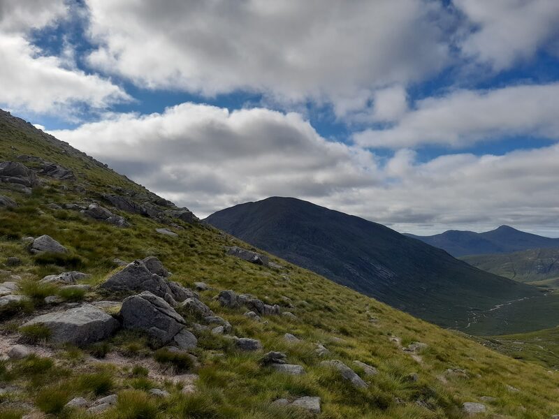 Beinn nan Aighenan whole, a broad dome in cloud shadow across a glen, seen from a slope of granite boulders, with a braided burn below and far peaks beyond.