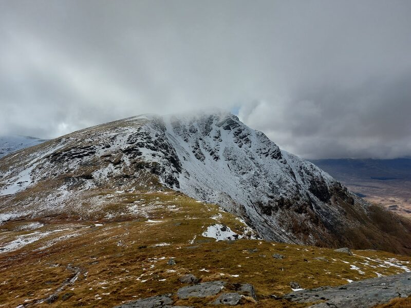 The steep, craggy end of Beinn Achaladair under a dusting of snow, its top in cloud, seen from the grassy ridge beside it.