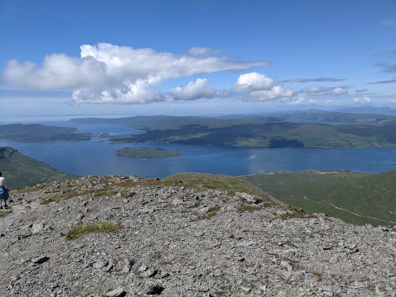 The sea loch and its islands far below the stony upper slopes of Ben More, under a blue sky, with a walker small at the left edge.