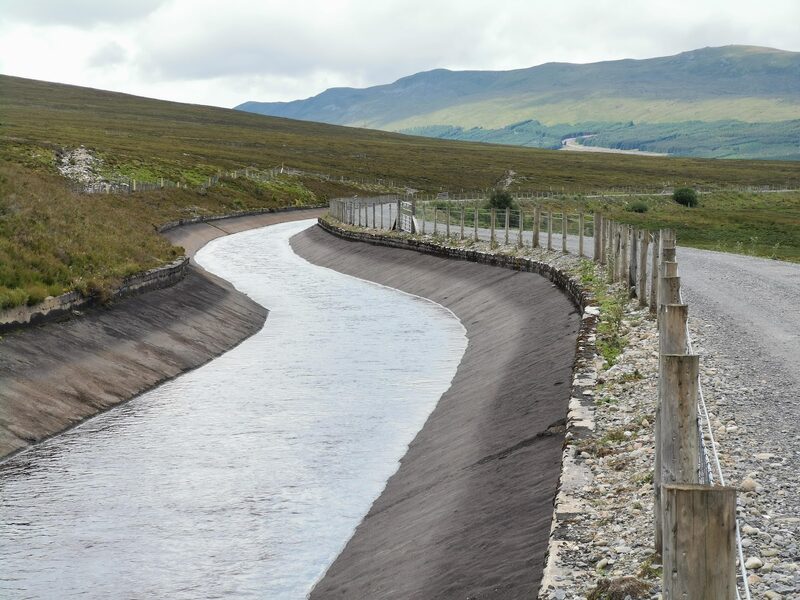 The concrete aqueduct full of water, a fence and the gravel track beside it, hills beyond.