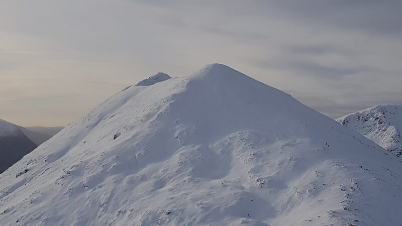 A cone of a summit under deep snow, with a snowy hill beyond.