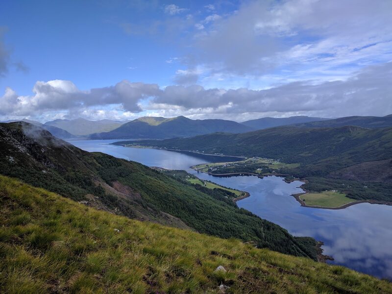 Looking down from a grassy slope high on the mountain to a sea loch and its narrows, with hills beyond under broken cloud.