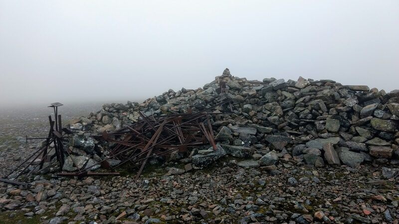 The big summit cairn in mist, rusted iron fence posts leaning in front of it.