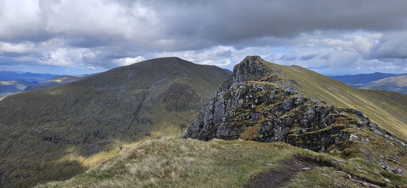 A rocky tor on the grassy ridge of Sgùrr a' Chaorachain, hills beyond under broken cloud.