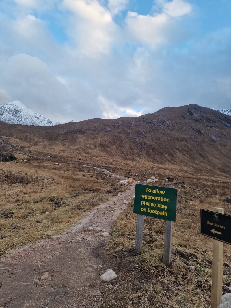 The path over brown moor with a green sign asking walkers to stay on the footpath and a fingerpost reading Ben Starav, a snow-covered top far to the left.