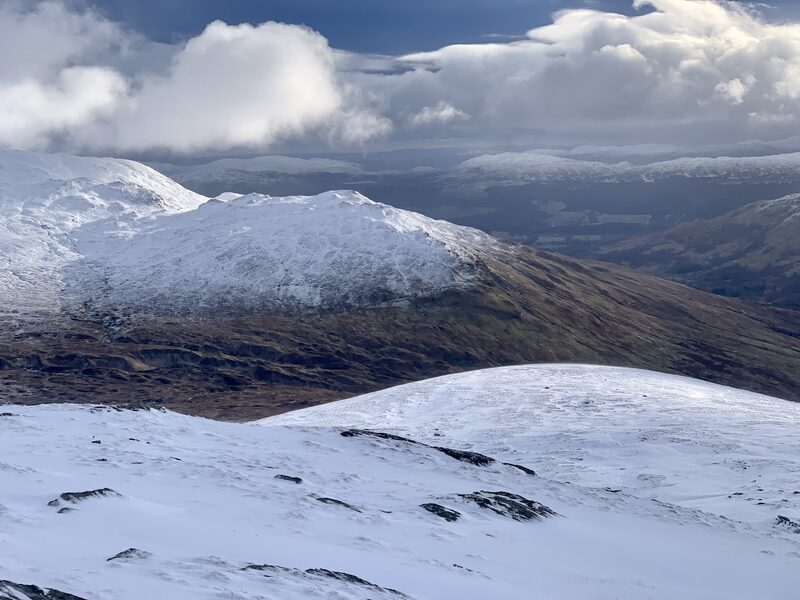 From snow-covered slopes, a snow-capped hill across a brown glen with banks of cloud and far white tops beyond.