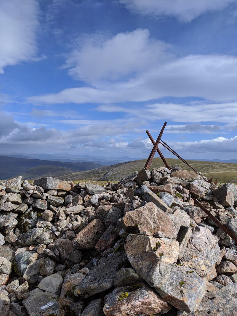 The summit cairn close to, two rusted iron posts crossed above the stones, with blue sky and far hills.