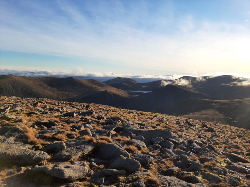 The bouldery top in low sun, hills and a far loch beyond with cloud lying in the glens.
