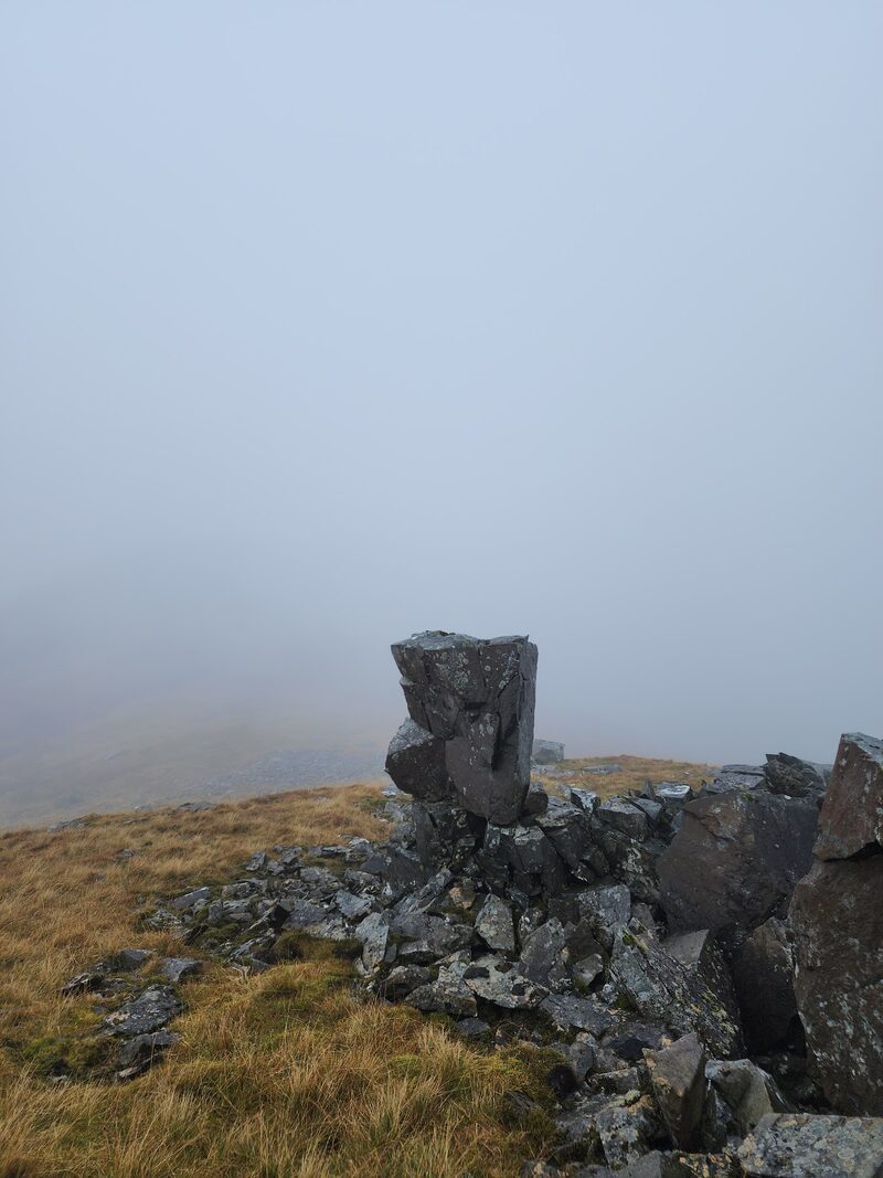 A leaning block of dark rock on a stony top in thick mist.