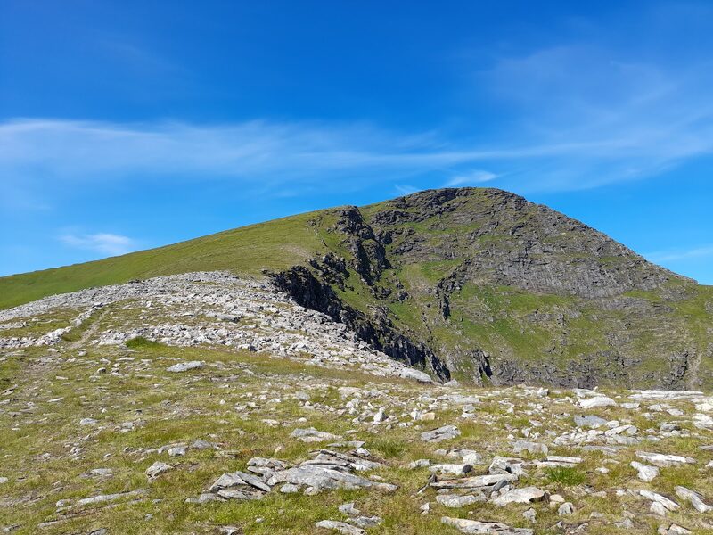 The crags and summit from near, across a stony shoulder of the ridge under a blue sky.