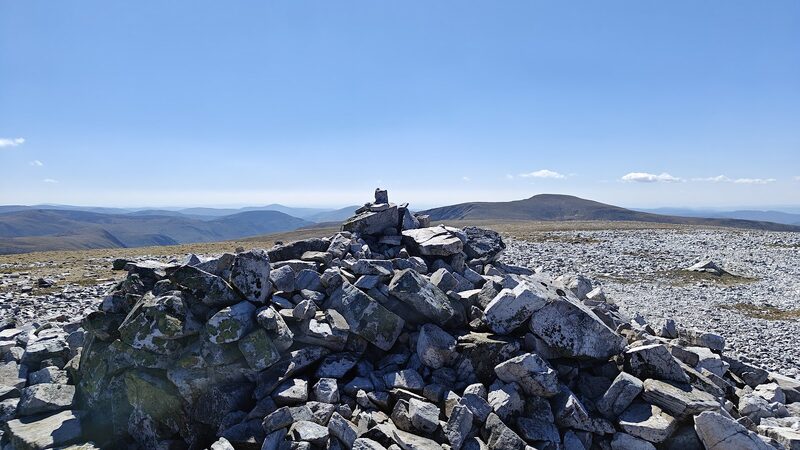 The summit cairn, a large pile of lichened blocks on a flat top of pale gravel, with distant hills under a clear sky.