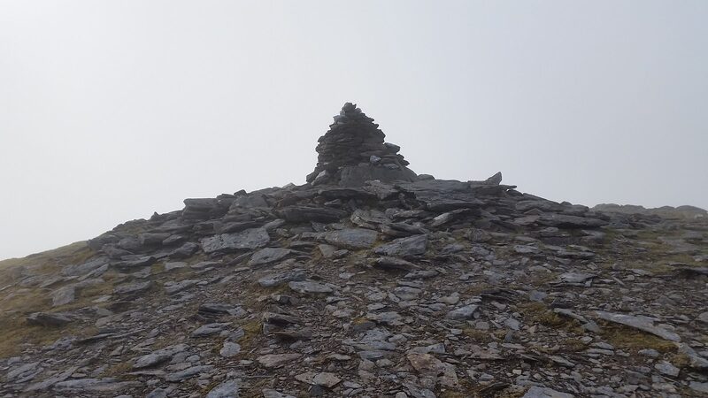 A tall cairn of flat stones on a stony top in thick mist.
