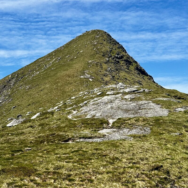 A steep green ridge rising to a pointed top on Stob Coir' an Albannaich, under a blue sky.