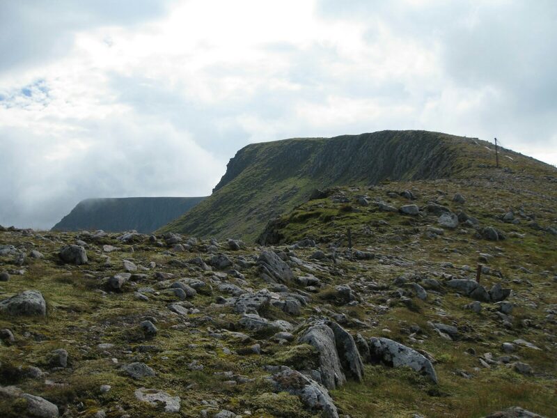 A flat-topped shoulder of the hill edged with crags, seen across bouldery ground under a heavy sky.