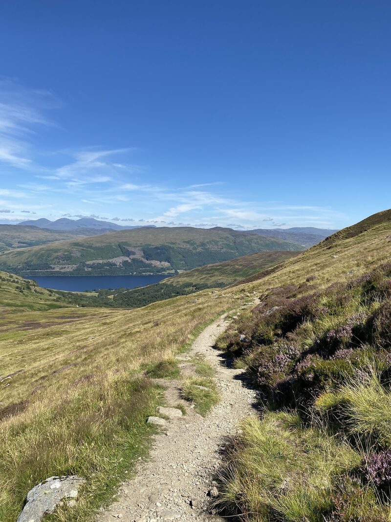 A gravel path through heather on the lower hill with the loch and its far shore below under a blue sky.
