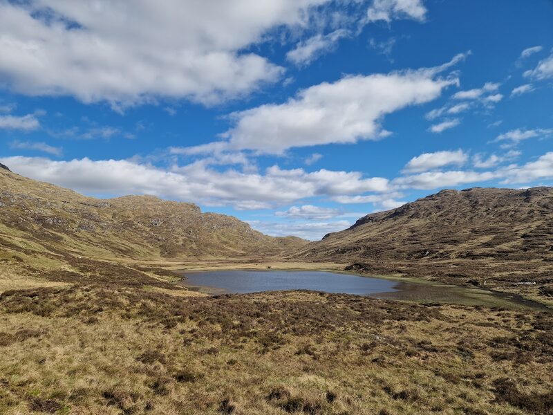 A hill loch in a hollow of heather and tawny grass, craggy slopes rising on either side under blue sky and white cloud.