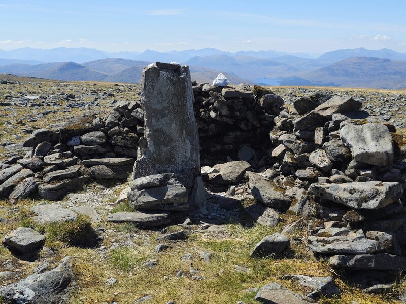 The summit's trig pillar inside its low ring of stones, far blue hills behind.
