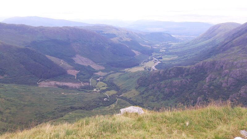 The view from the hillside down a long green glen, its road, fields and woods far below.