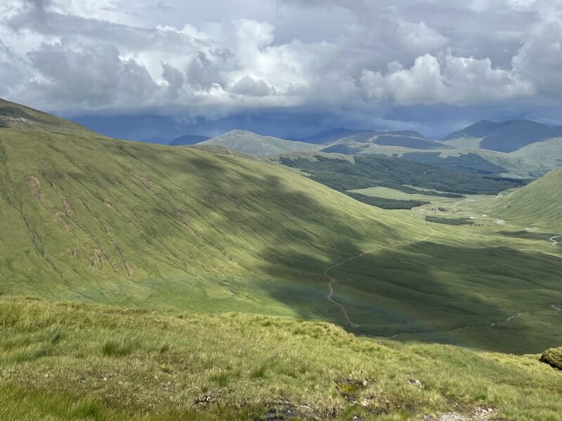 The view down from a high grass slope into a green glen with a winding track, shower clouds over the hills beyond.
