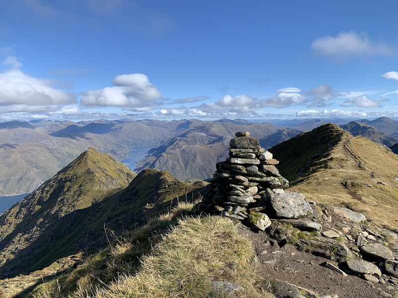A cairn of stacked stones on a narrow grassy crest, with pointed tops and a sea loch beyond.