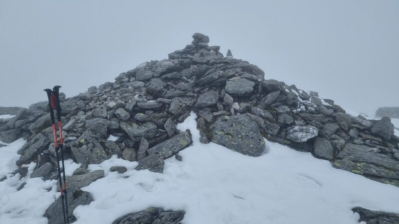 The large stone cairn on the top of Ben More in snow and mist, a pair of walking poles propped against it.