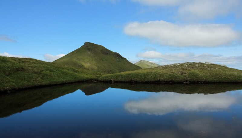 A steep green cone with a level top reflected in a still hill lochan, a second summit just showing behind it.