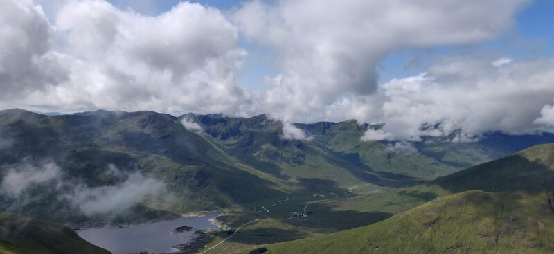 A loch on a glen floor far below green slopes, with heavy cloud on the hills across it.