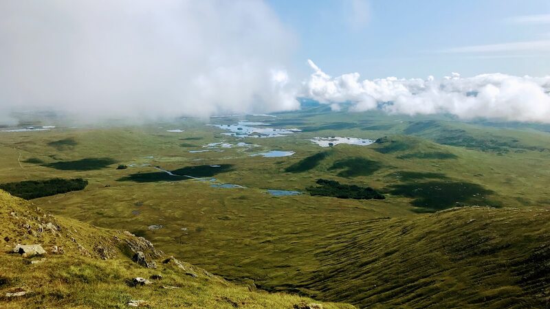 From the hill's grassy slopes, the wide moor and its scatter of lochans under a bank of cloud.