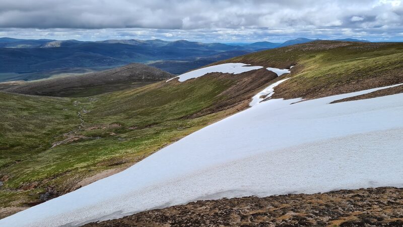 The view down from the hill over a long snow patch to green slopes and blue hills.
