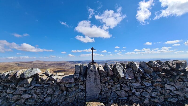 The drystone wall on the top of Creag Leacach with an old iron post standing above it against a blue sky.
