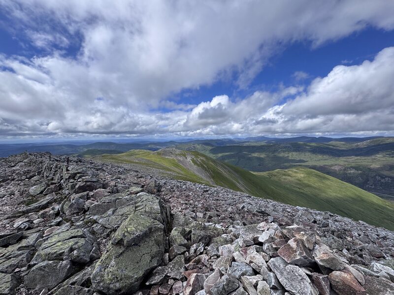 The top of Creag Leacach: tumbled pinkish slabs and the stub of a drystone wall, a green ridge running away below under big white clouds.