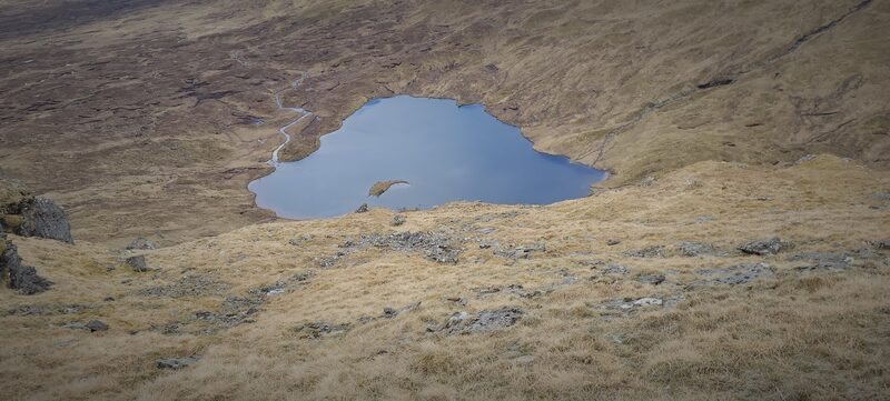 A blue hill loch in a hollow of tawny grass and peat, seen from the slope above.