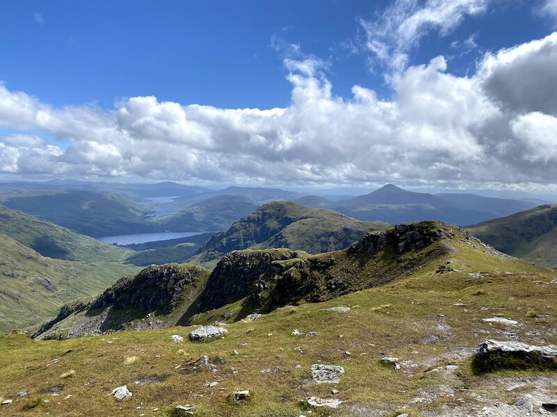 The view from high on Beinn Ìme: stony grass in front, craggy tops below, a loch and a pointed hill far beyond under white cloud.
