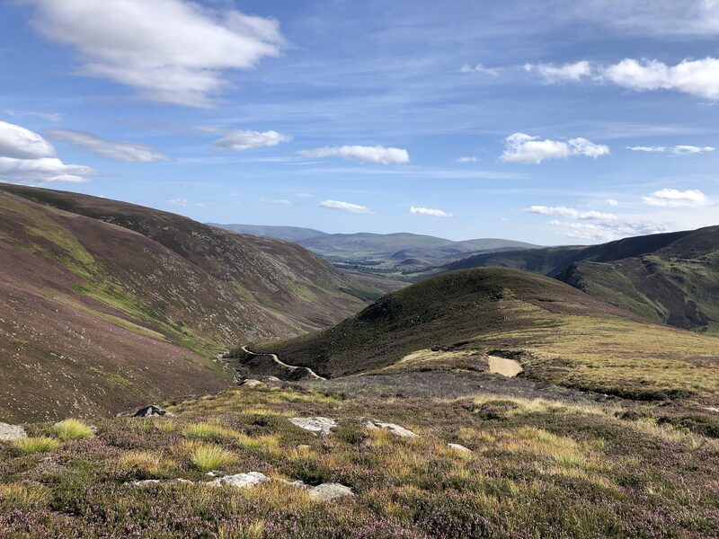 The view from the heather slopes of Mount Keen down a deep glen to far hills under a blue sky.