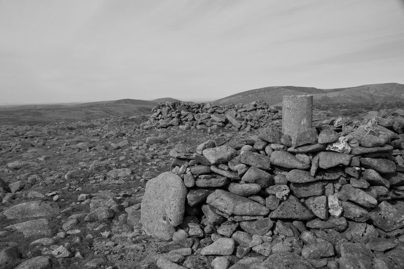 The summit in black and white, a trig pillar inside a drystone shelter wall on a field of stones.