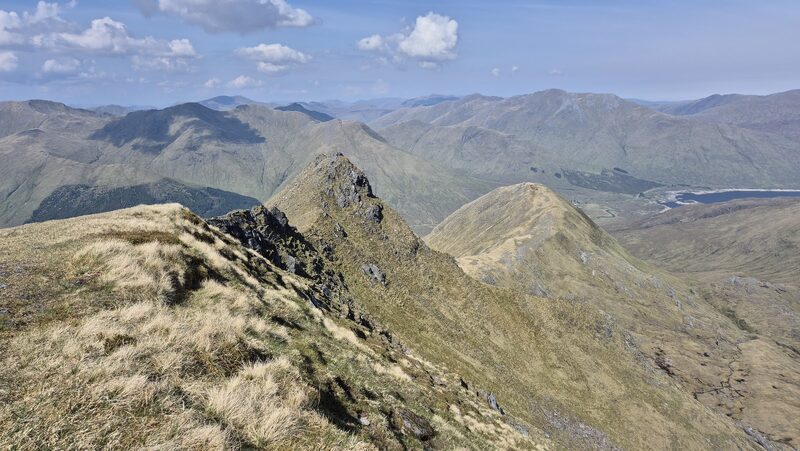 A grassy top looking along a ridge to a rocky peak, with ranks of hills and a loch beyond.