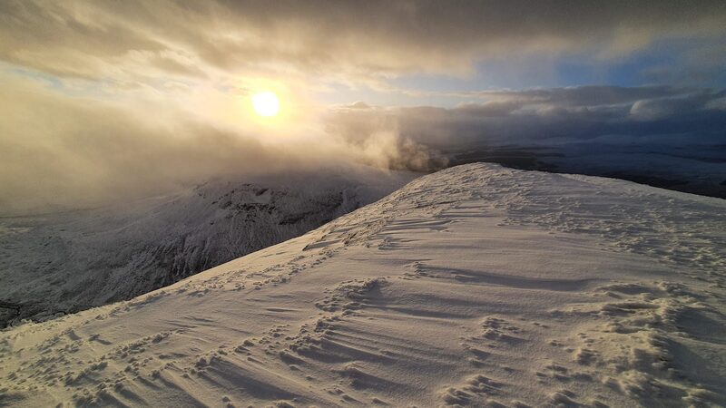 The broad ridge of Ben Wyvis under wind-carved snow, low sun breaking through cloud over the corrie.