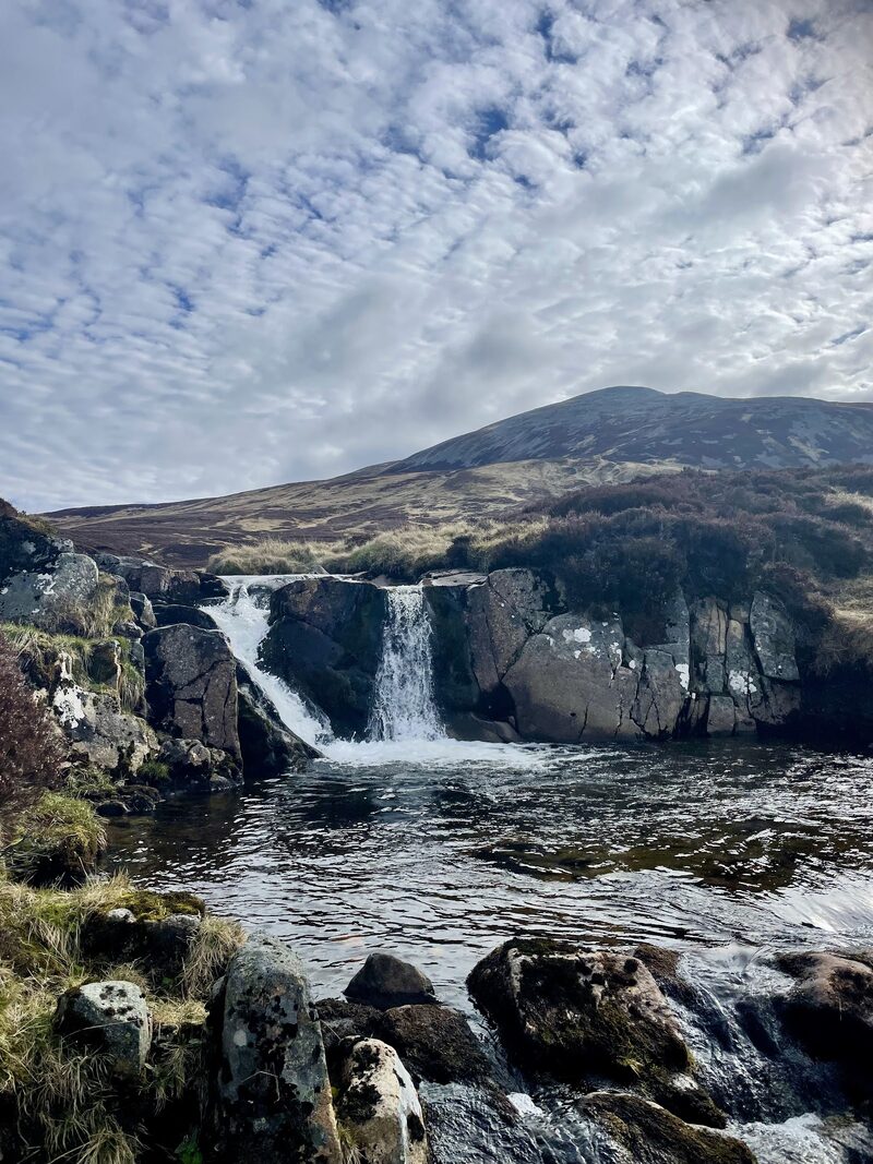 A waterfall dropping into a dark pool on the burn, with the stony dome of Càrn an Tuirc above under a dappled sky.