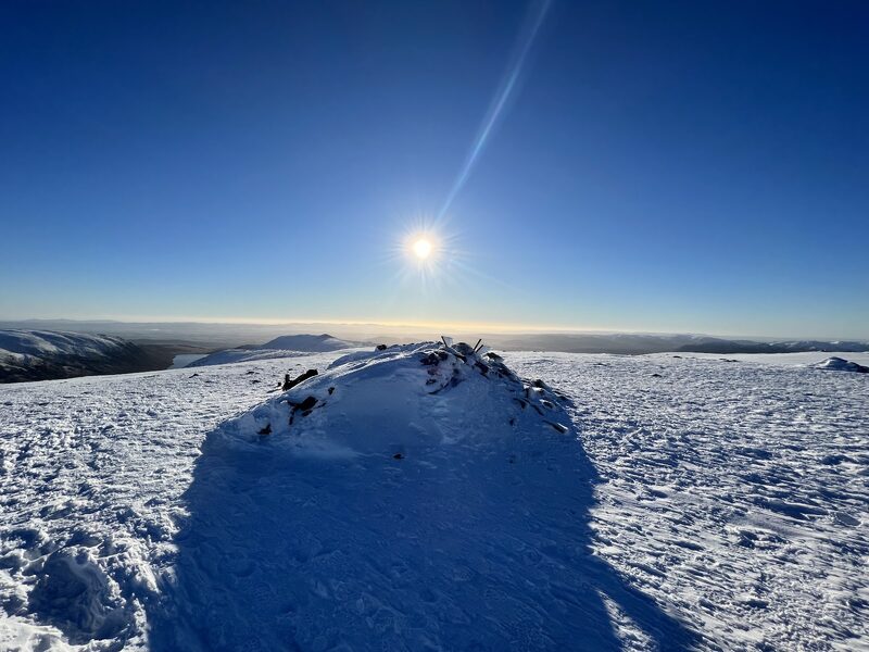 The snow-covered summit cairn of Ben Chonzie under a low sun, a reservoir in the glen far below.
