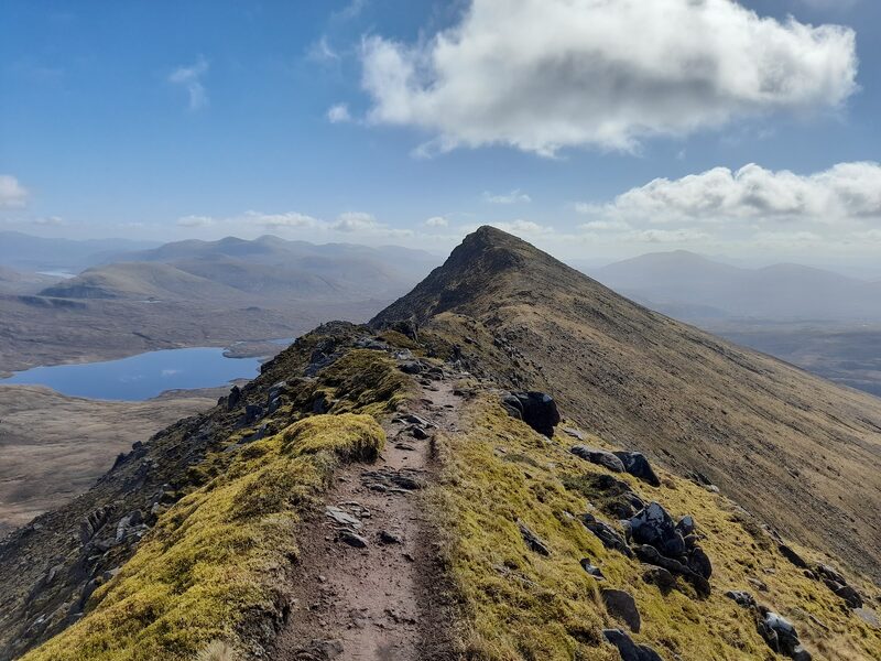 A worn path along a narrow grassy ridge high on Slioch, running to a pointed top with a loch far below.