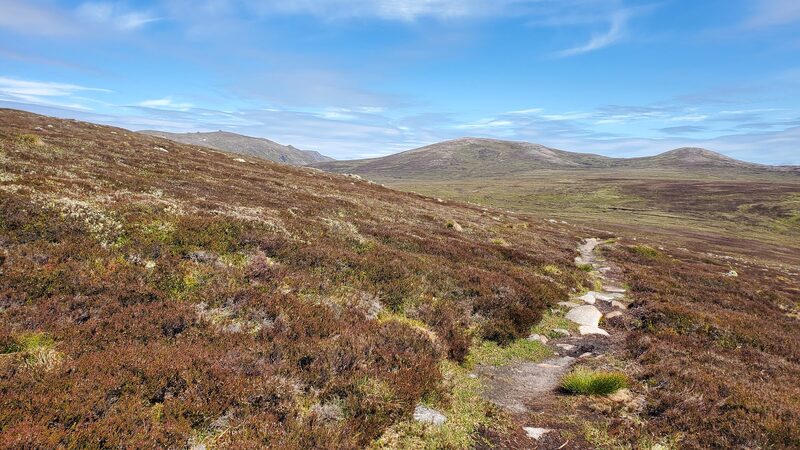 A path through heather towards the rounded tops of Beinn Bhreac under a blue sky.