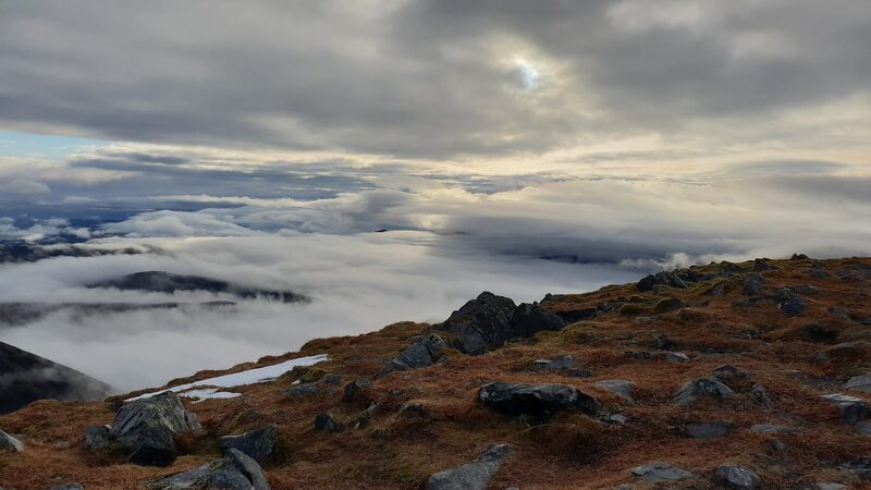 Boulders and russet moss on the summit above a sea of cloud, a low sun behind grey sky.