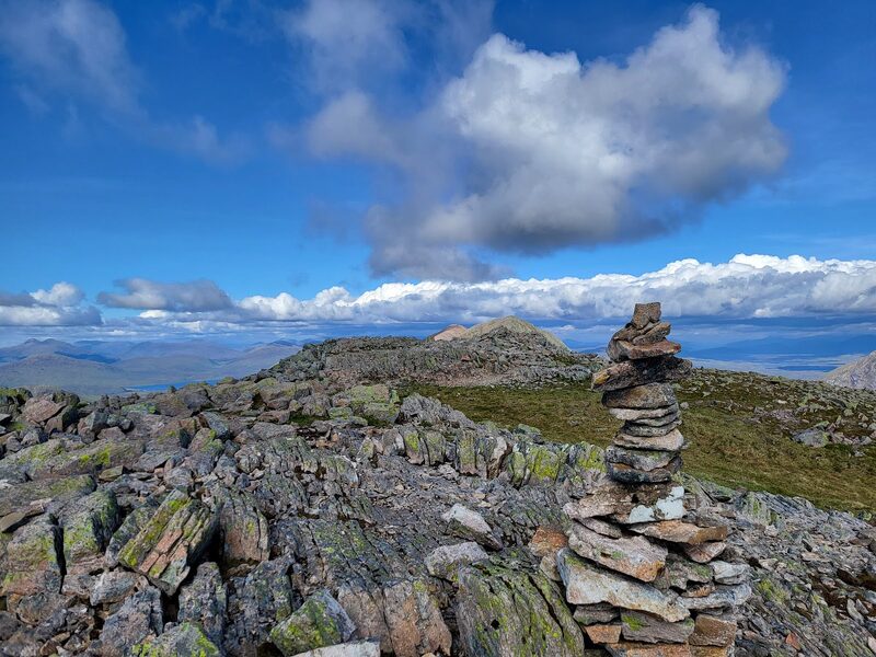 The cairn among lichened boulders, with the mountain's ridge running on to its further tops under a tall cloud.