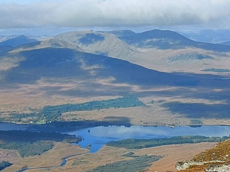 The view down from high ground to a loch among pines and a wide moor, cloud shadows moving across it.