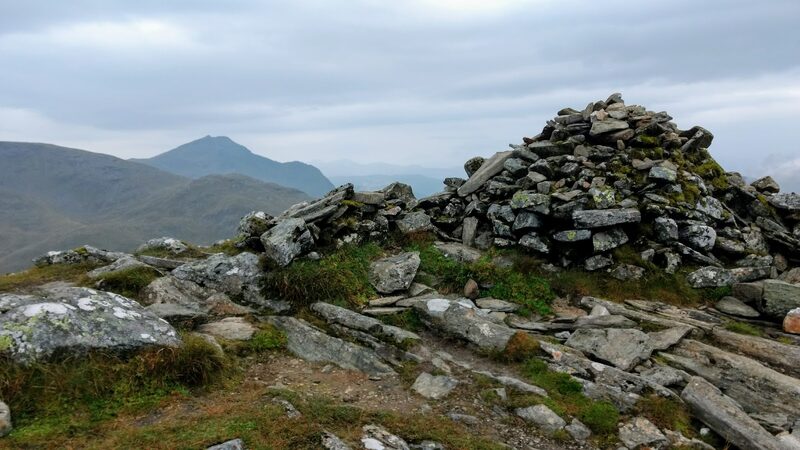 The mossy summit cairn of grey stones on Beinn Dubhchraig, with a sharp peak far beyond a rounded hill under grey cloud.