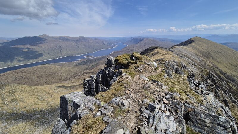 A rocky crest of tilted slabs along a ridge, with a long loch in the glen below and a rounded top ahead.