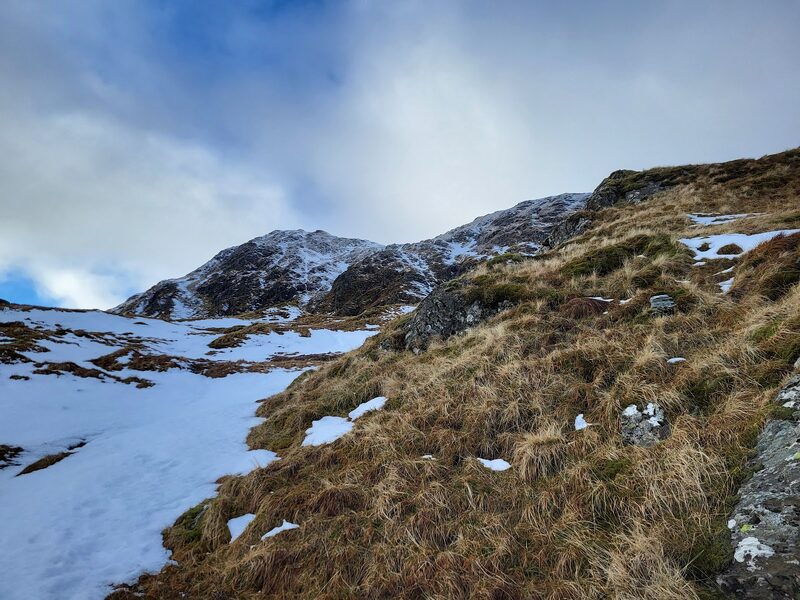Rough grass and rock under patches of old snow, with a rocky top ahead under cloud.
