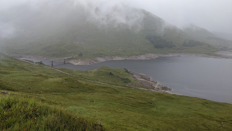 A road bridge crossing an arm of a loch below a grassy hillside, with the hills beyond in mist.